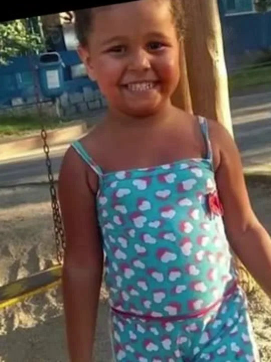 Ana Clara de pé num parquinho, de macacão estampado com corações, sorrindo.