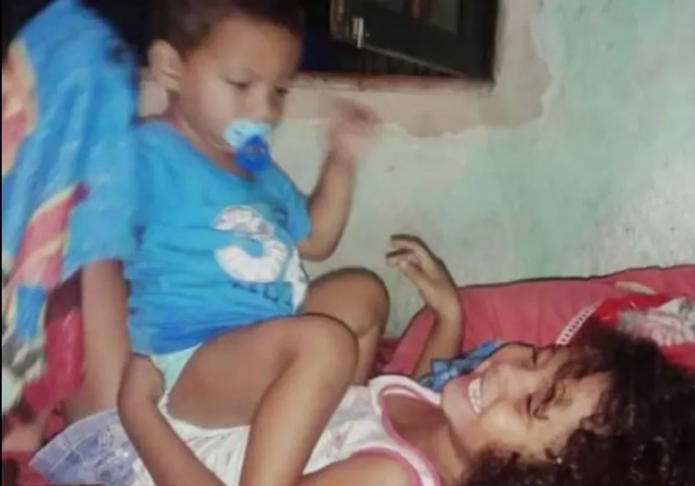Foto antiga da família: Ana Clara deitada numa cama, rindo, ao lado do irmão pequeno de chupeta.